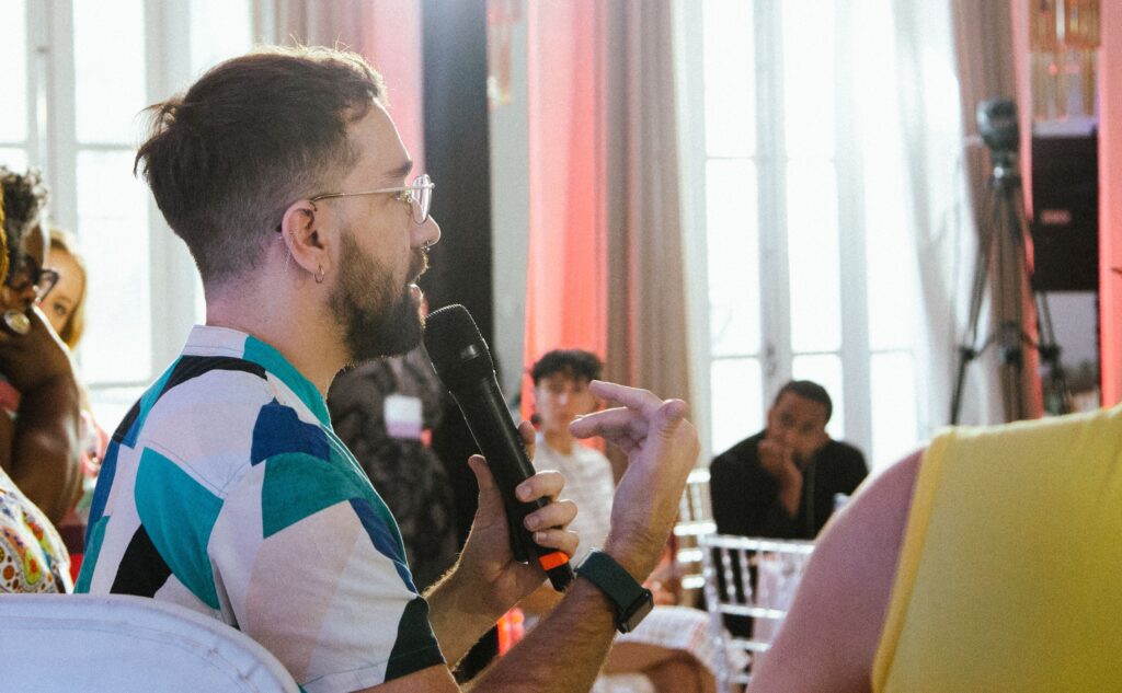 A side profile of Ivens Reyner speaking into a black microphone during an event. He is wearing glasses and a white shirt with a geometric pattern in teal and dark tones. In the blurred background, other attendees are seated in a bright room with large windows and pinkish curtains.