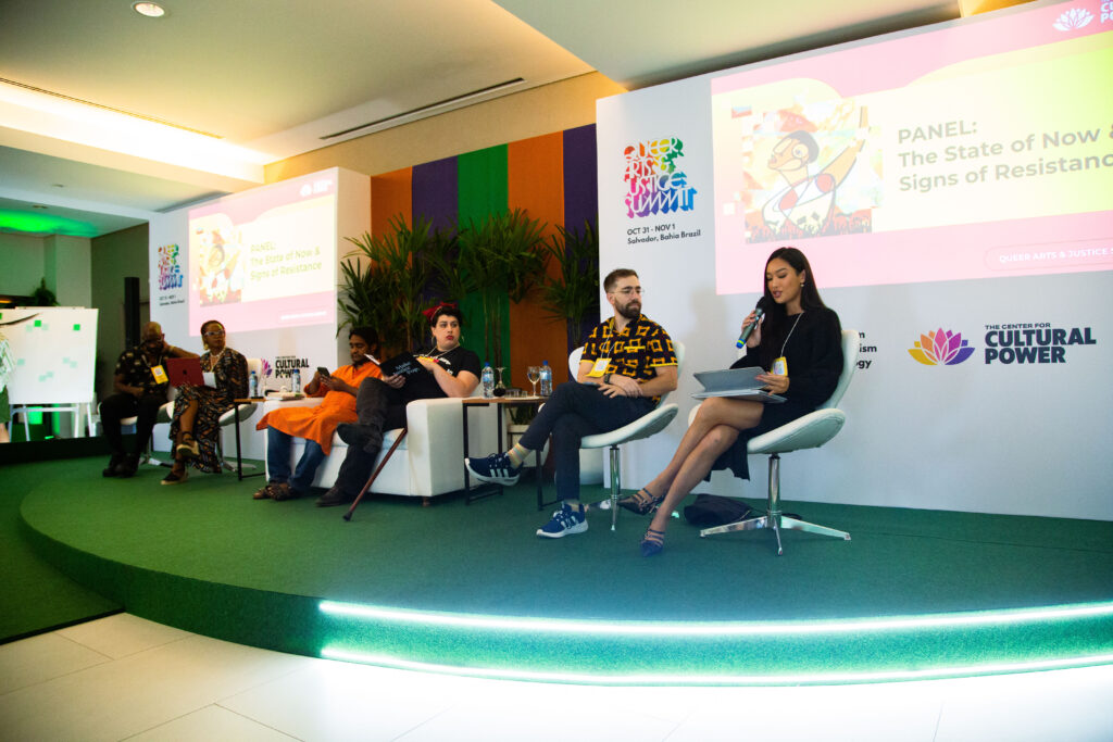 A wide shot of a stage at a conference titled "The State of Now & Signs of Resistance." Ivens Reyner is seated on a white modern chair as part of a diverse six-person panel. He is wearing a black shirt with a yellow geometric pattern and dark trousers. To his right, a woman in a black dress is speaking into a microphone. The stage has a green carpet, and the background features a large screen with the event's branding and "The Center for Cultural Power" logo.
