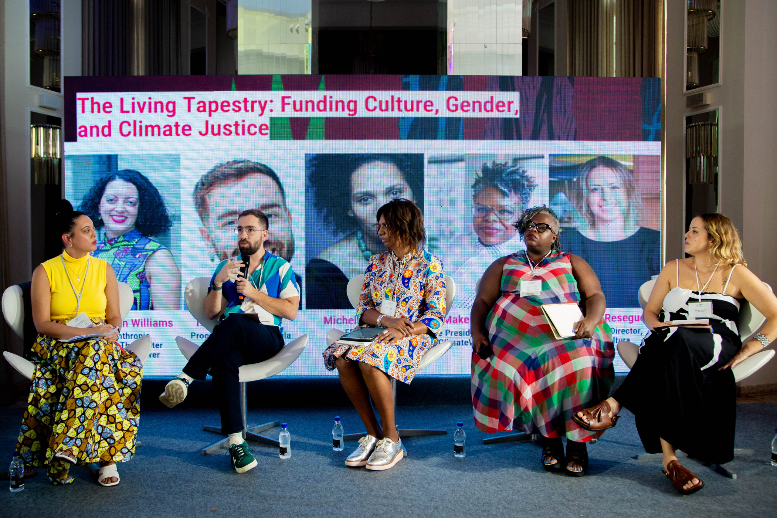 A panel discussion featuring five diverse speakers sitting in white modern chairs on a stage. Behind them is a large digital screen displaying their portraits and names under the title o the event. The speakers, four women and one men of various ethnicities, are dressed in colorful, professional attire. One man in a teal and white shirt is speaking into a microphone while the others listen attentively.