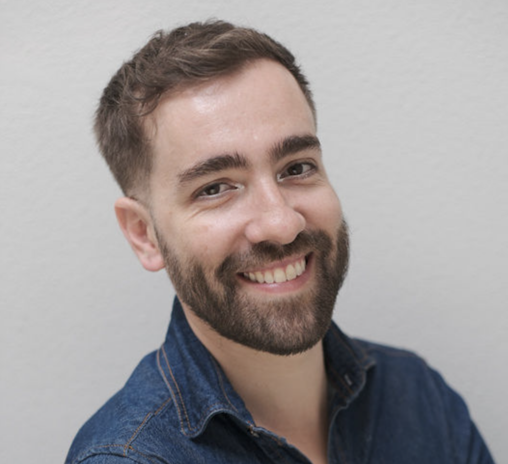 A portrait of Ivens Reyner, a man with short brown hair and a groomed beard, smiling warmly at the camera. He is wearing a dark blue denim shirt and is positioned against a neutral, light-gray textured background.